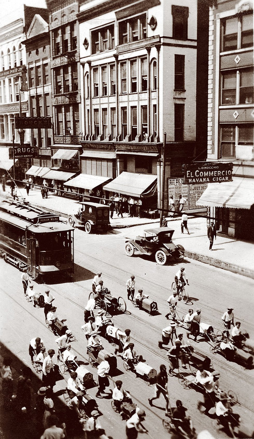 A parade of soapbox derby cars, pushed from behind, makes its way down Main Street in front of a trolley. 